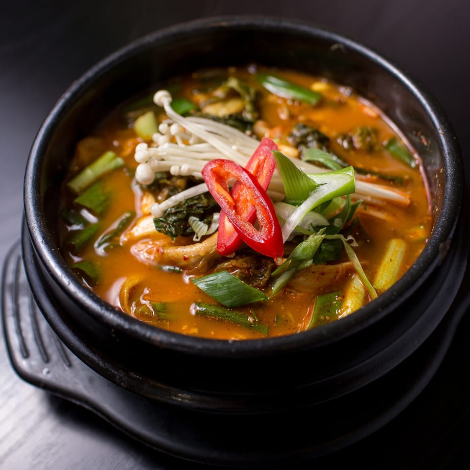 Close-up of a vibrant orange stew in a black bowl, garnished with mushrooms, scallions, and red chili slices.
