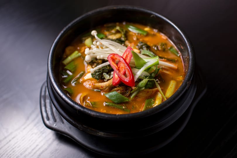 Close-up of a vibrant orange stew in a black bowl, garnished with mushrooms, scallions, and red chili slices.