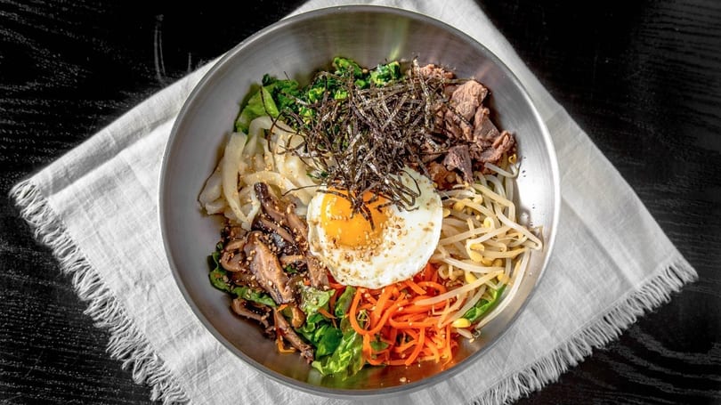 A colorful bibimbap bowl with beef, vegetables, and a fried egg, on a white napkin.