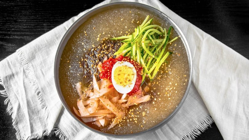 Korean cold noodles (naengmyeon) with cucumber, egg, radish, and red sauce in a metal bowl.